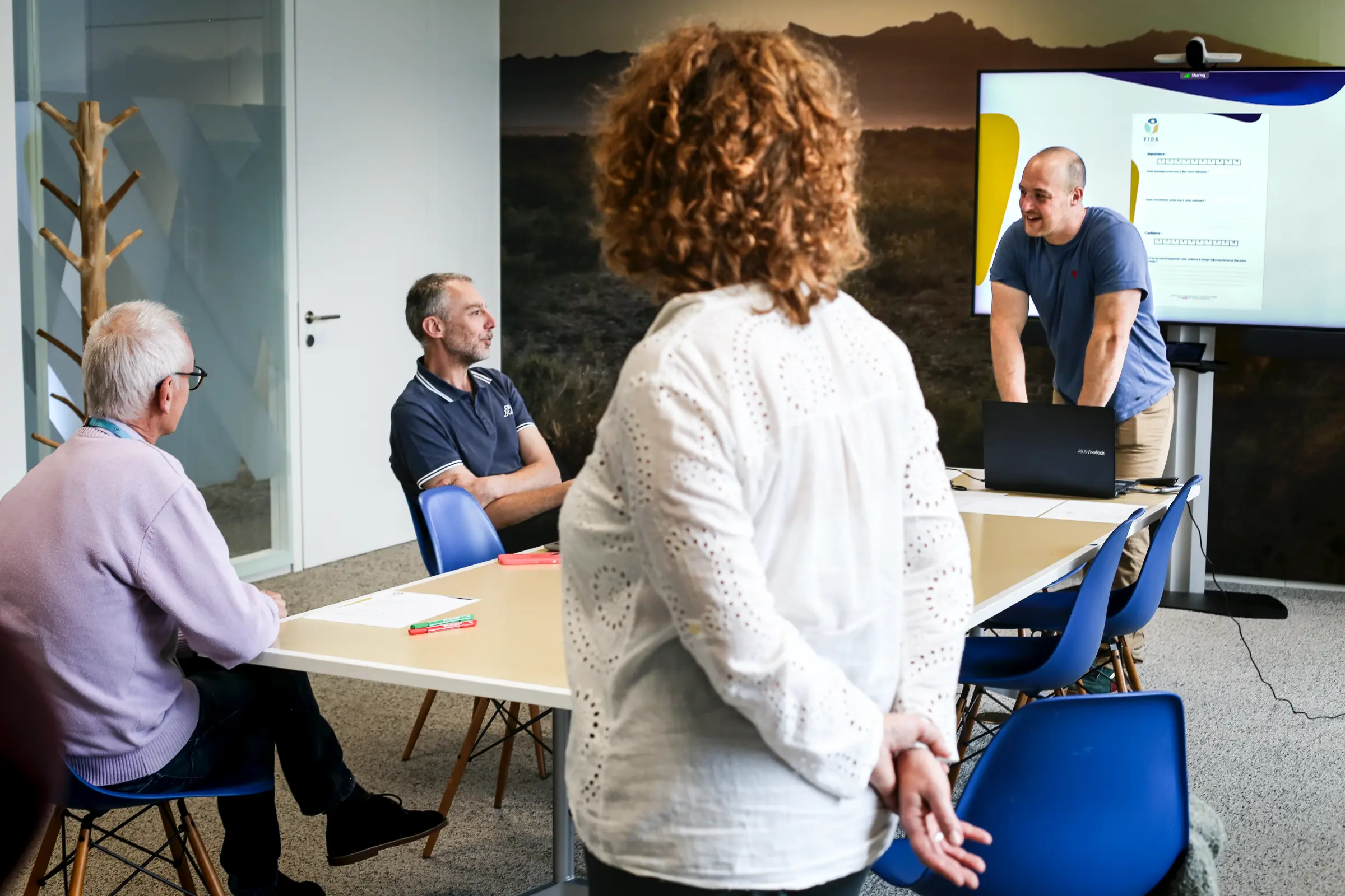 Salle de formation VIDA Consult: un animateur présente à un groupe d’adultes assis autour d’une table, écran affichant une présentation.
