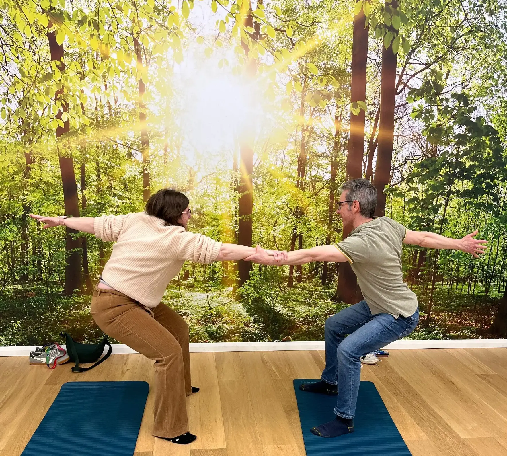Deux personnes en train de faire des exercices de squats sur tapis de yoga bleu, dans une pièce avec un mur imprimé forêt lumineuse.